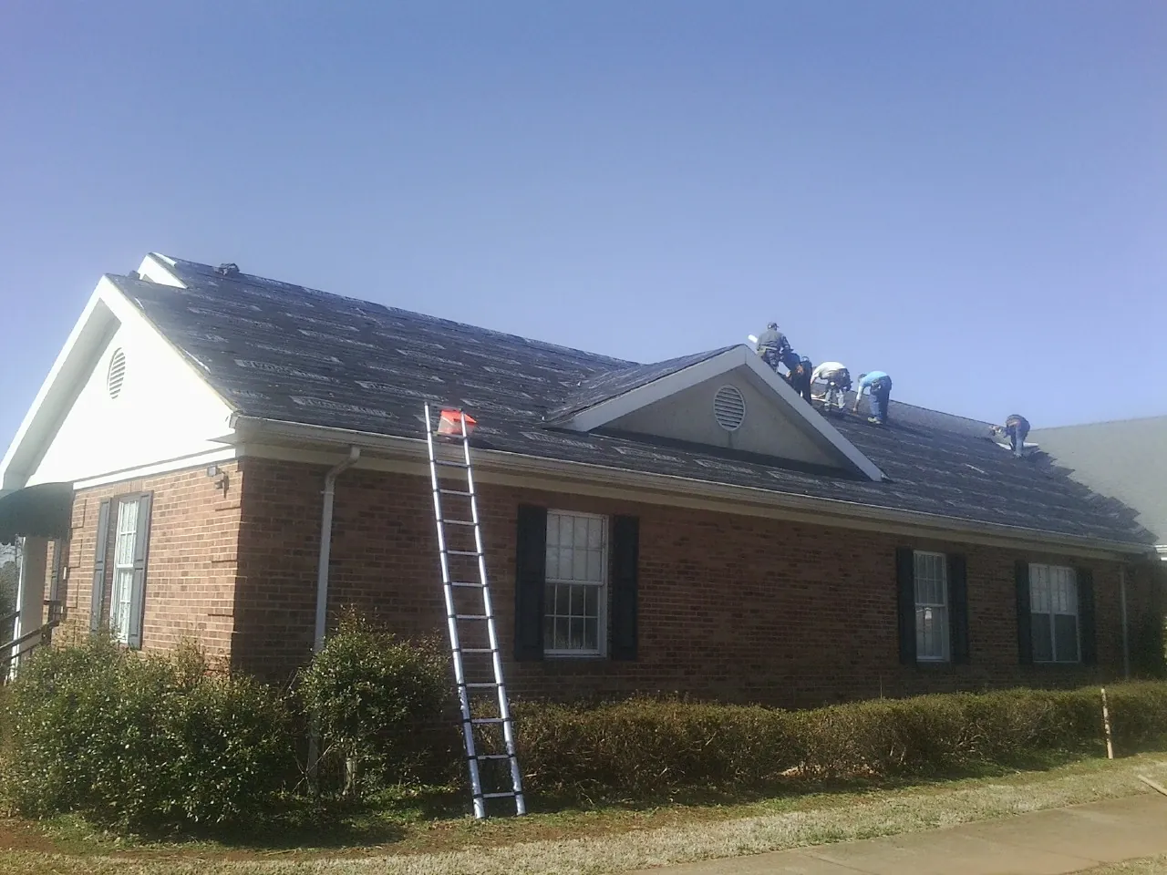 Crew tearing shingles off a brick ranch roof with a ladder set — Metal Roof Repair work by our crew in Falconer
