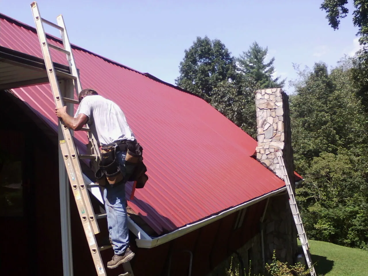 Roofer on a ladder beside a red metal roof and stone chimney — Soffit & Fascia Repair by Roofing Brothers in Falconer